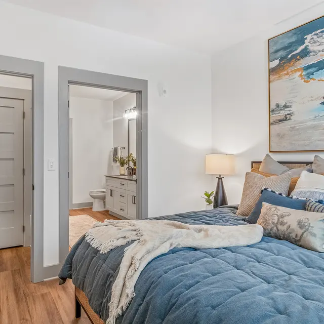 A modern bedroom featuring a large bed with blue bedding and multiple decorative pillows. There is a bedside lamp and a piece of artwork on the wall. A doorway to a bathroom is visible in the background.