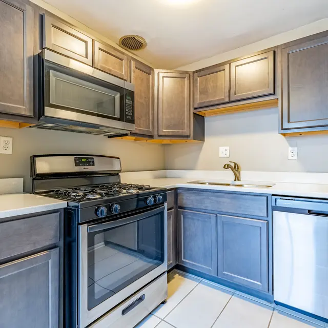 A modern kitchen featuring dark wood cabinets, stainless steel appliances, and a white countertop. The space includes a gas stove, microwave, and dishwasher.