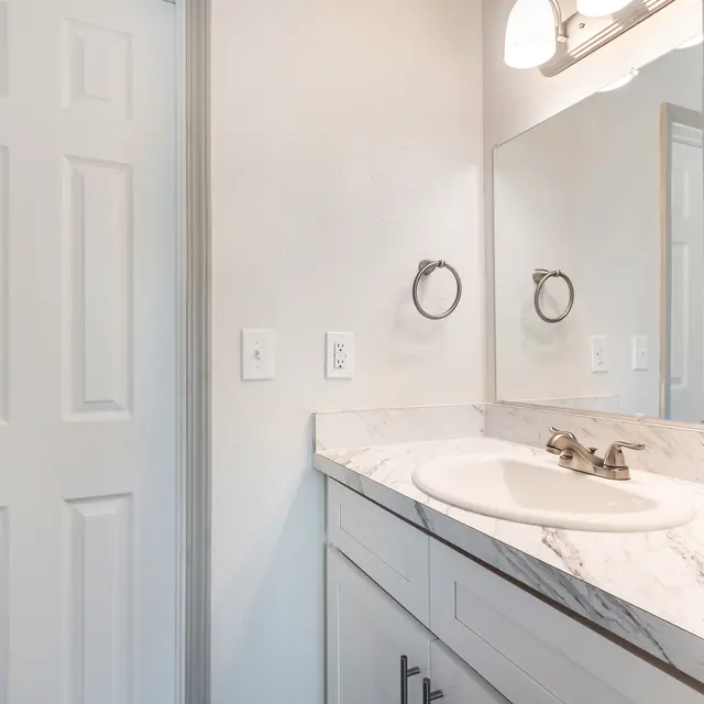 A modern bathroom with a marble countertop, a sink, a large mirror, and a door on the left.