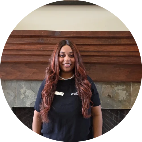 A woman with long, wavy hair is smiling while standing in front of a fireplace. She is wearing a black shirt with a name tag and a pearl necklace.