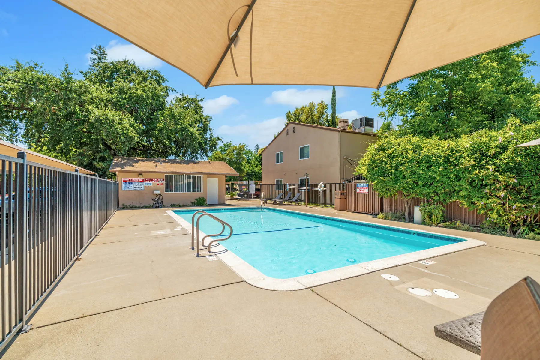 A clean, inviting swimming pool area surrounded by trees and a fence, with a shaded seating area nearby.