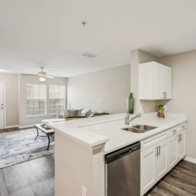 A view of a modern kitchen and living room area in an apartment, showing a white countertop, stainless steel appliances, a ceiling fan, and a cozy seating area with natural light.