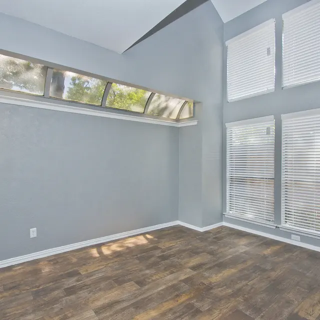 A spacious room with grey walls and a wood-style floor, featuring large windows with blinds, allowing natural light to illuminate the space.