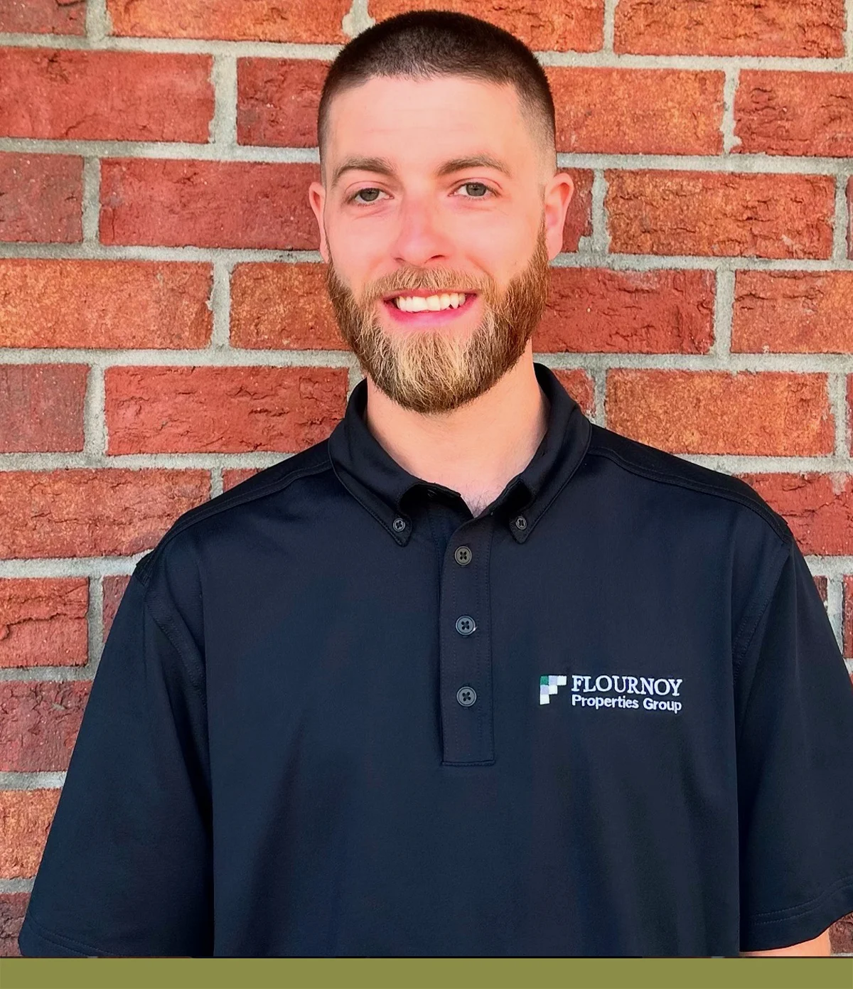 Professional Portrait Against Brick Wall A smiling man with a beard, wearing a black polo shirt with a logo, stands in front of a brick wall. The background showcases a colorful arrangement of red bricks.