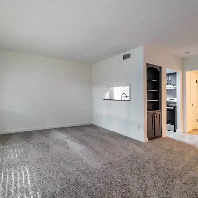 A spacious, empty living room with light gray carpet and bright natural light coming through large windows. There is an opening to a kitchen area with modern cabinets visible in the background.