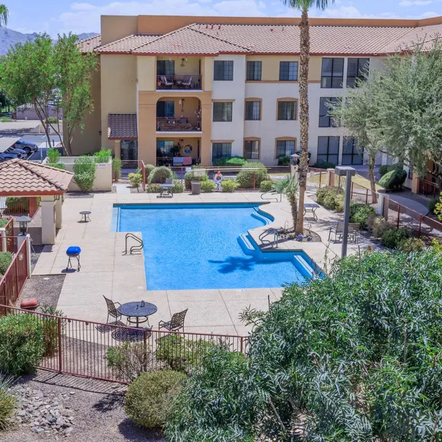 Resort Pool Area A view of a resort-style swimming pool surrounded by landscaped grounds, with palm trees and lounge chairs. The building in the background has multiple stories with balconies overlooking the pool area.
