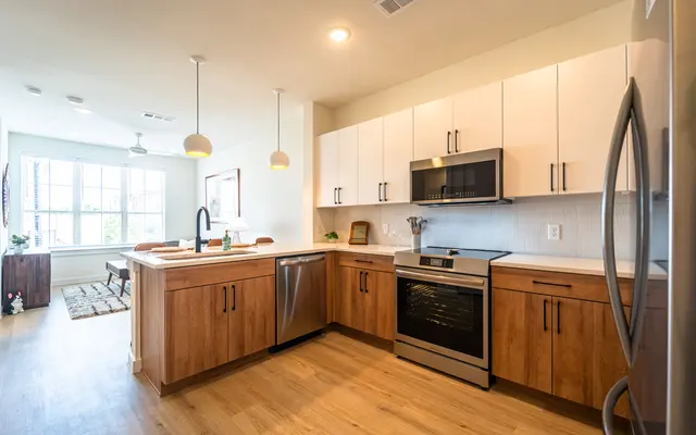 A modern kitchen with wood cabinetry, stainless steel appliances, and a light-colored countertop. A seating area is visible in the background with a rug and window.