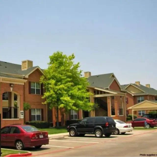 A residential apartment complex featuring red brick buildings and green landscaping, with parked cars near the entrance.