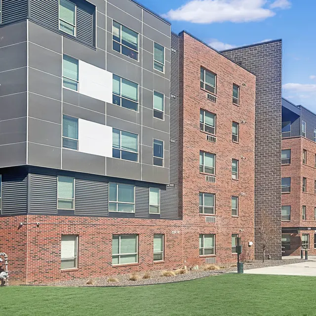 A modern apartment building featuring a combination of brick and gray paneling. The structure has multiple windows and a grassy area in the foreground with a sidewalk.