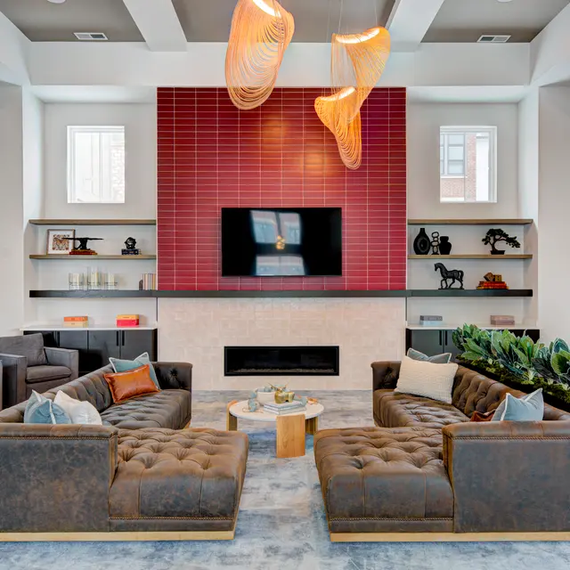 A modern living room featuring comfortable sofa seating, a decorative coffee table, and a wall-mounted television above a stylish fireplace. The backdrop is characterized by red tile and shelves with decorative items, complemented by unique light fixtures hanging from the ceiling.