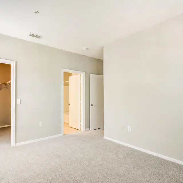 An empty bedroom featuring beige carpet and light gray walls. There are two doorways: one leading to a closet and another to an adjacent space. The overall space is bright and airy.