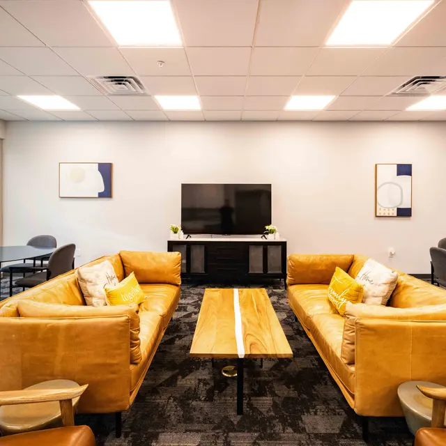 A modern meeting space featuring two yellow sofas facing a wooden coffee table. A large TV is mounted on the wall, surrounded by minimalistic artwork. There are several tables with chairs in the background and large windows letting in natural light.