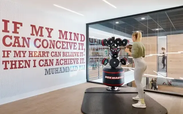 Motivational Gym Quote by Muhammad Ali A woman training in a modern gym featuring a punching bag and a motivational quote on the wall.