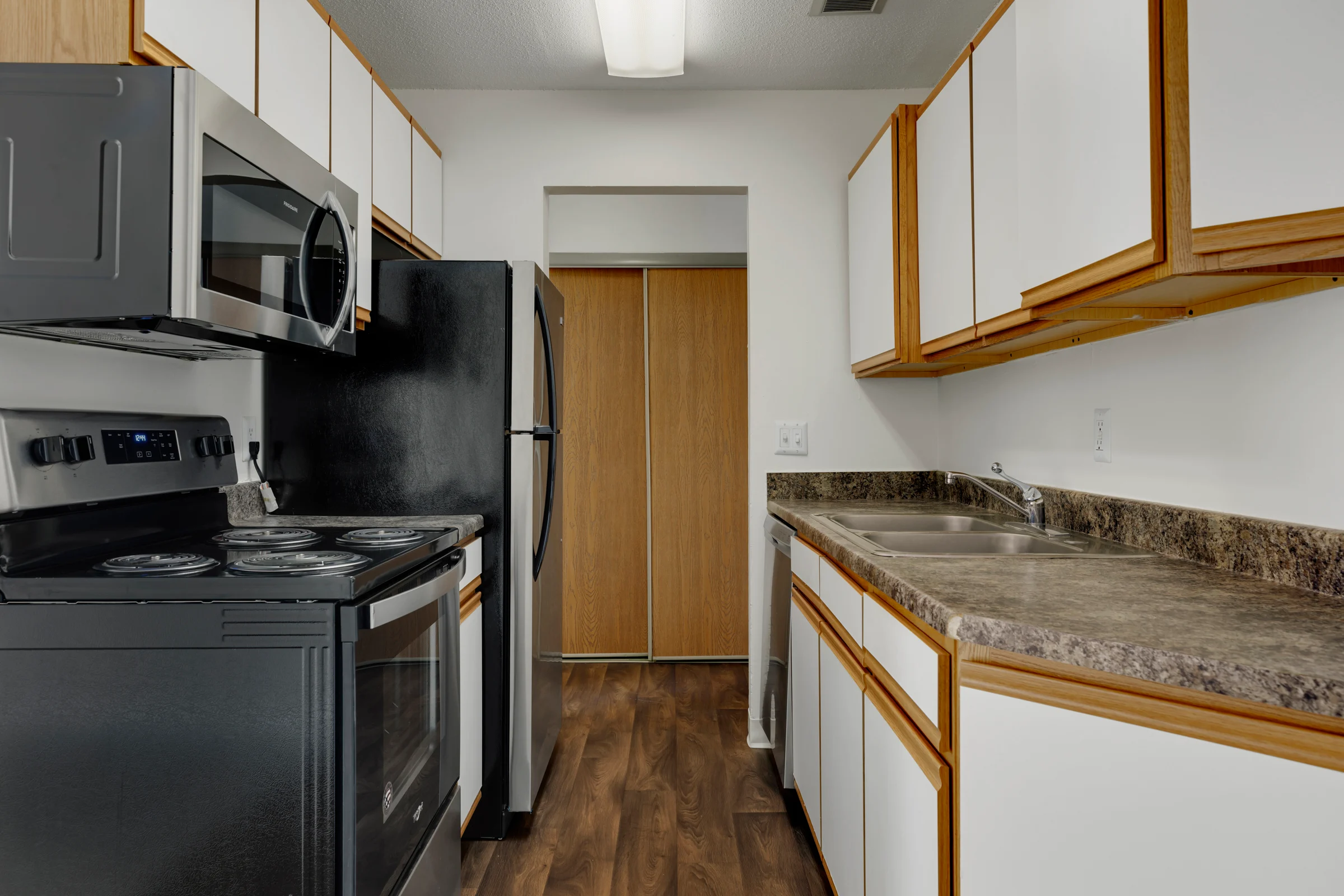 A modern kitchen featuring a black stove and microwave, a black refrigerator, and light wood cabinets with a granite countertop. The space includes a sink and a doorway leading to another room.