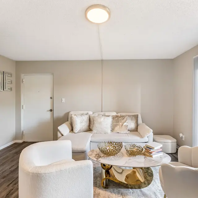 A modern living room featuring a white sofa with decorative pillows, two white armchairs, and a round coffee table with decorative items. The room has a light grey color scheme and large windows letting in natural light.