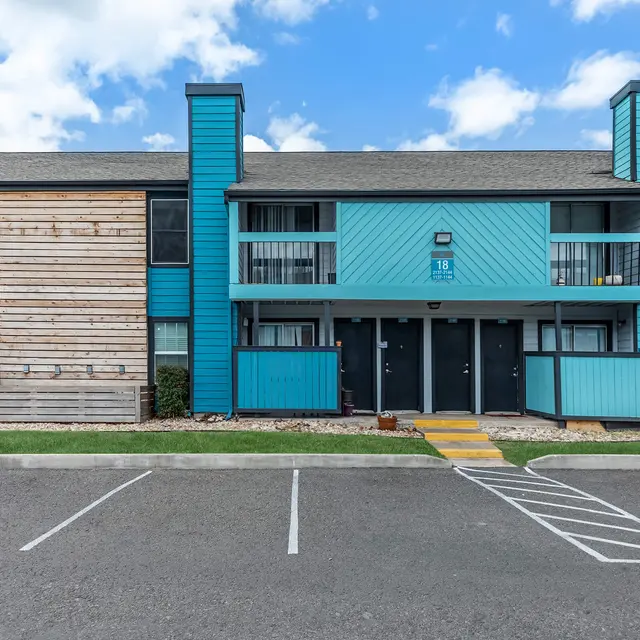 Exterior view of an apartment complex featuring a mix of blue and wooden paneling, with balconies, in a parking lot under a blue sky.