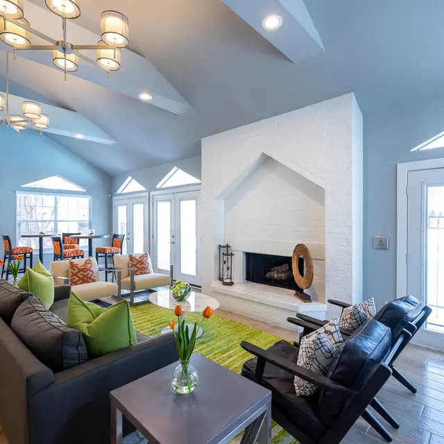 Bright and Modern Living Room Design A bright and modern living room with large windows, featuring a gray sofa with colorful pillows, a coffee table, and two black chairs. A contemporary chandelier hangs from the ceiling, and a white fireplace is the focal point of the room, alongside green area rugs. Several chairs and a table are visible in the background, creating a spacious and inviting atmosphere.