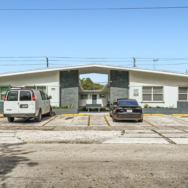 A modern apartment complex with a unique architectural design, featuring a symmetrical front and a parking lot in the foreground. The building is primarily white with gray accents, and several cars are parked in designated spots.