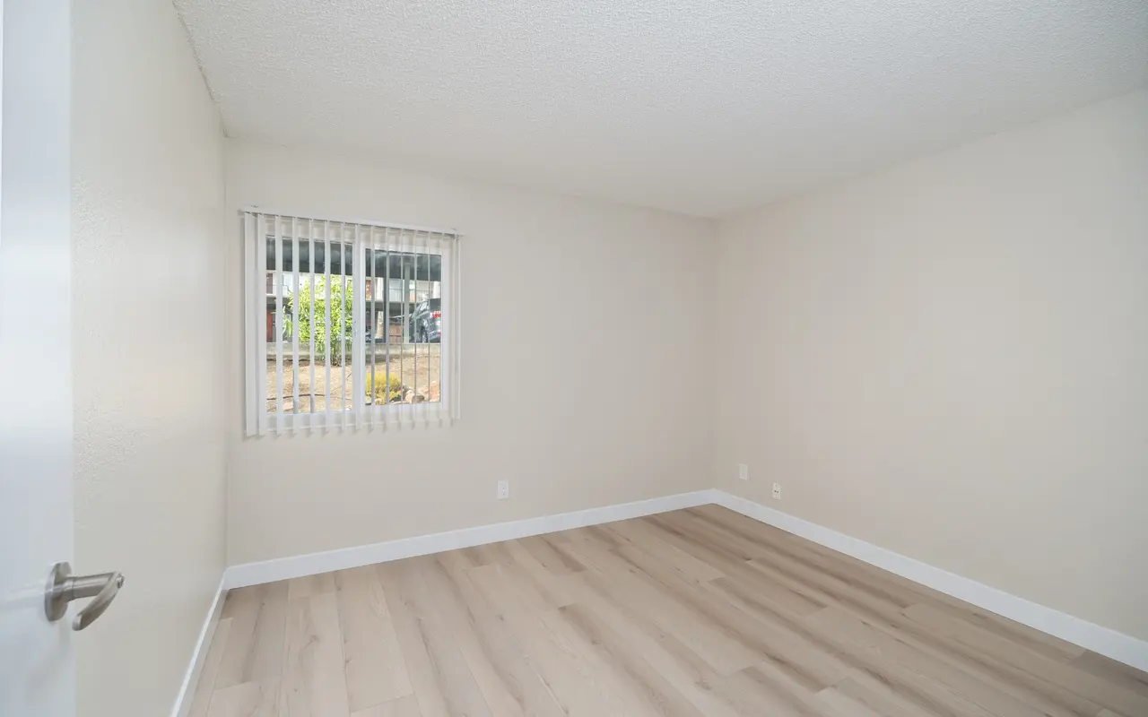 An empty room with light-colored walls and wooden flooring, featuring a window with blinds.