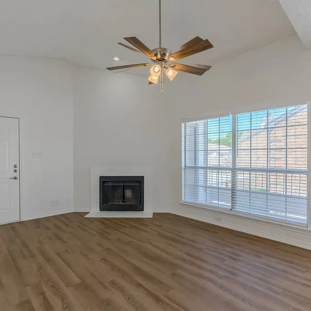 A spacious living room featuring a ceiling fan, a fireplace, and large windows that allow natural light, along with light-colored walls and wooden flooring.