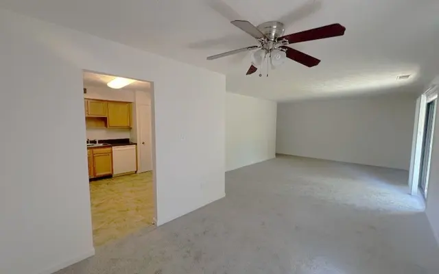 Empty Living Room with Kitchen Access A spacious empty living room with a ceiling fan and light-colored carpet. An open doorway leads to a kitchen area with light wood cabinets and an adjacent doorway. The walls are painted white, and the room has natural light coming in from the adjacent area.