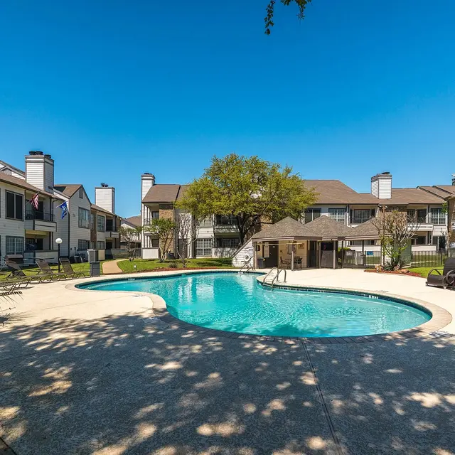 A sunny day at a residential pool area featuring a curved swimming pool surrounded by lounging chairs, manicured lawns, and apartment buildings in the background.