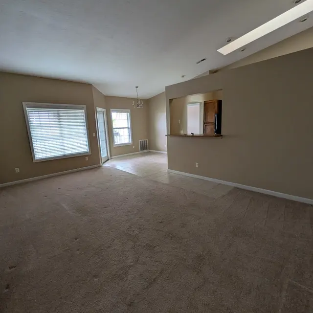 A spacious living room area with beige walls and carpet, featuring a window with blinds, a sliding glass door, and a small bar area connected to the kitchen.