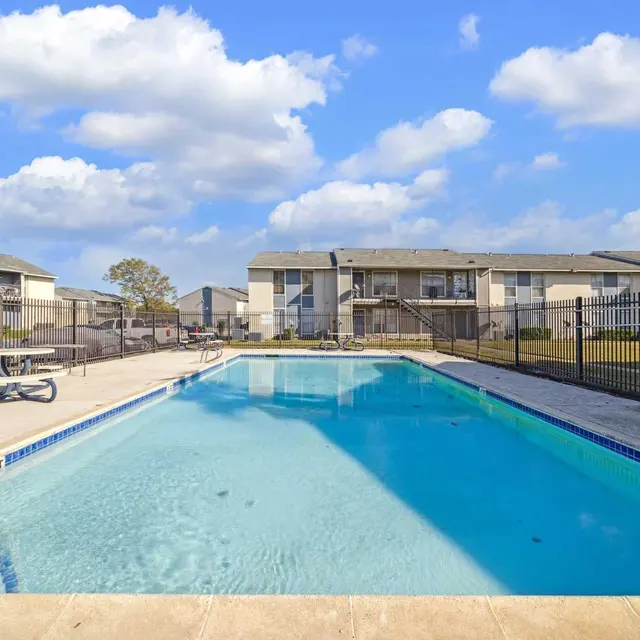 A clean, clear swimming pool surrounded by a fence and apartment buildings in the background under a blue sky with scattered clouds.
