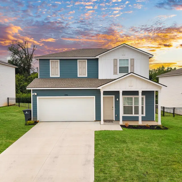 A modern two-story blue and white house with a well-maintained lawn and a double-car driveway during sunset. The sky is colorful with orange and purple hues.