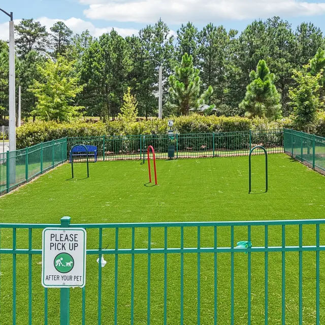 A small, fenced dog park featuring synthetic grass and a few agility equipment like hoops and jumps, surrounded by trees and a parking lot in the background.