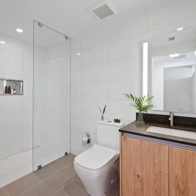 A stylish modern bathroom featuring a glass shower enclosure, a sleek toilet, and a wooden vanity with a dark countertop. The walls are adorned with white tiles, and a small plant adds a touch of greenery to the space.