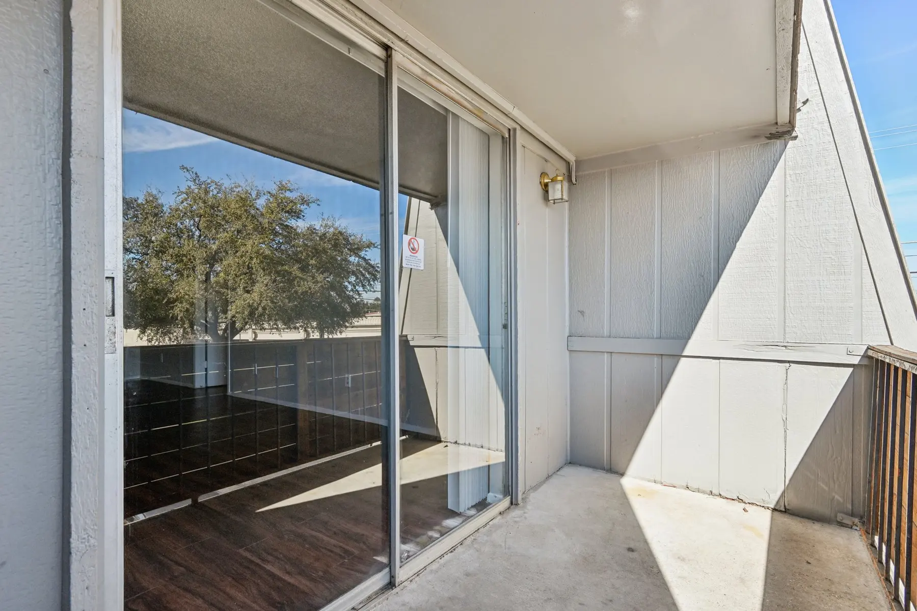 A balcony with sliding glass doors overlooking a sunny outdoor area with trees.