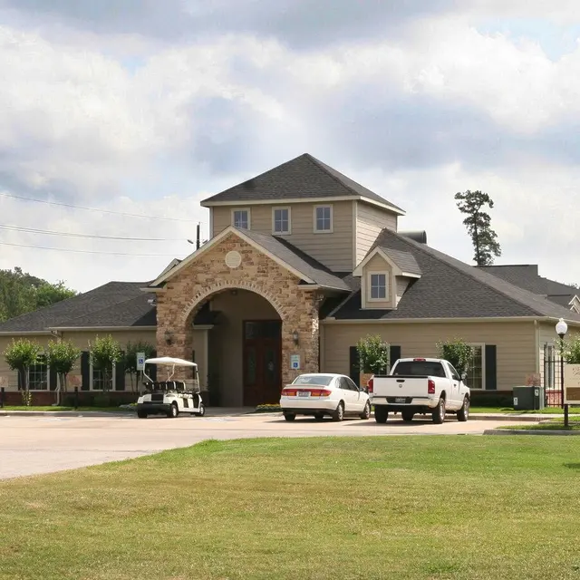 Modern Apartment Complex Front View A front view of a modern apartment complex with a stone and wood facade. There are three vehicles parked in the foreground, and the area is surrounded by green grass and small trees under a cloudy sky.