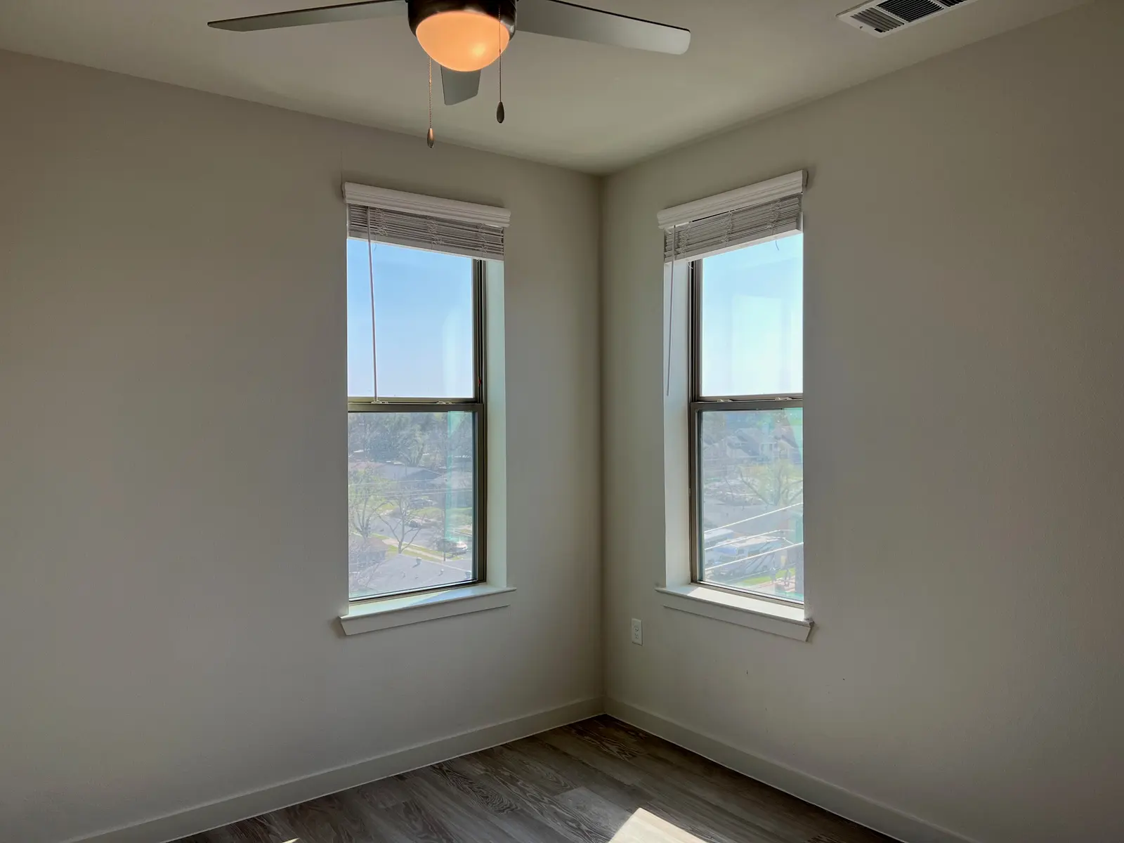 Bright Corner Room with Windows A well-lit corner room featuring two windows with blinds, clear blue skies visible through the glass. There is a ceiling fan with light omitted in the center of the room, and the floor is made of light-colored wood laminate.