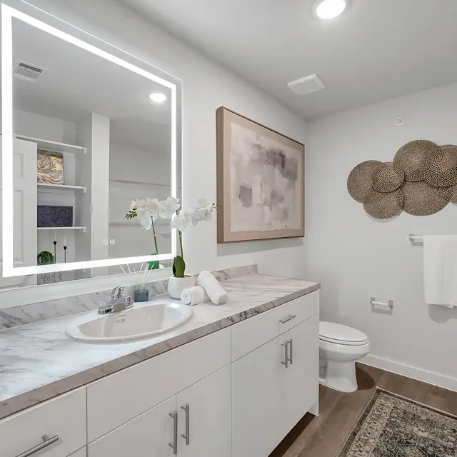 A modern bathroom featuring a sleek white vanity with a marble countertop, illuminated mirror, and a decorative wall piece made of woven materials. A toilet and shower curtain are visible, and the floor is finished in a wood-like texture.
