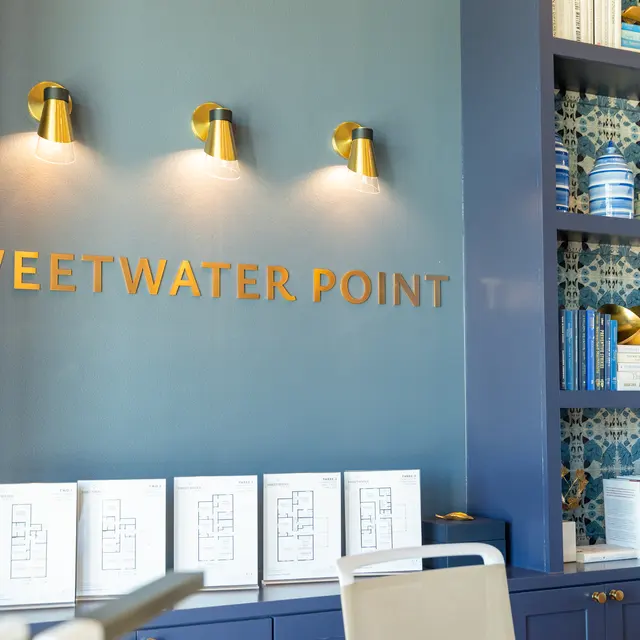 Interior view featuring a wall with the words 'SWEETWATER POINT' in gold lettering above illuminated wall sconces, alongside shelves filled with decorative items and books.