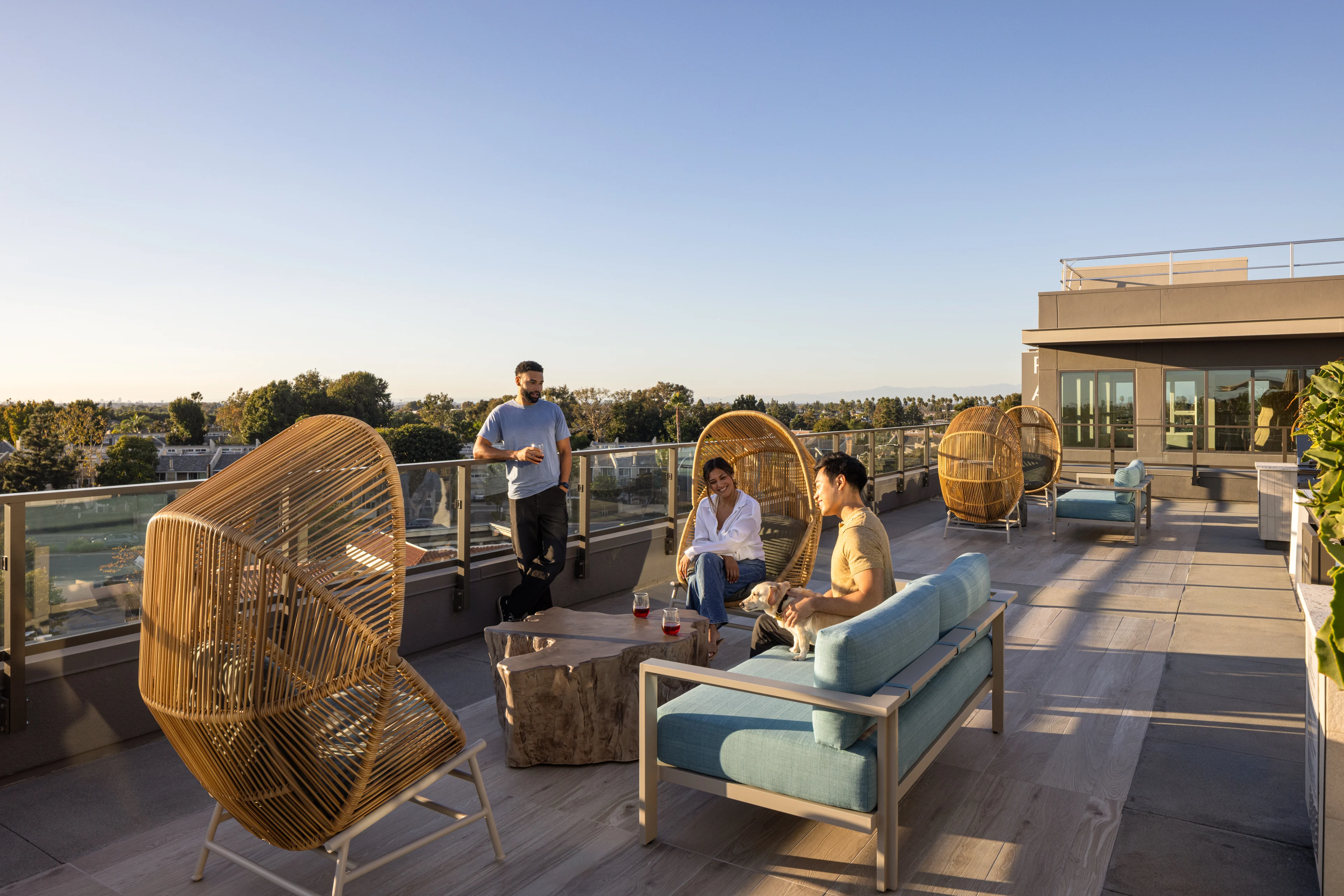 Rooftop Gathering at Sunset A rooftop area with three people socializing among modern furniture, featuring large woven chairs and a clear view of the sunset sky.