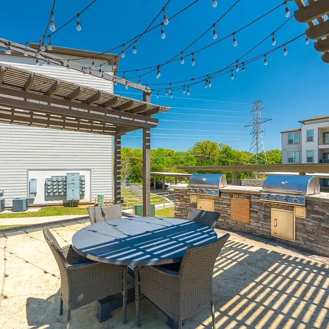 Outdoor dining and grilling area with a table and chairs, surrounded by string lights and apartment buildings in the background.