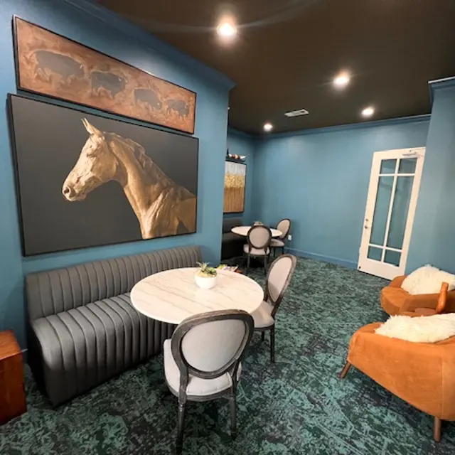 A stylish interior room featuring a blue wall, artwork of a horse, a circular table with a plant centerpiece, and various seating options including a gray banquette and orange armchairs.