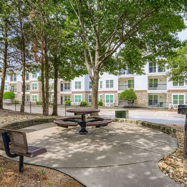 Outdoor Gathering Space A small outdoor gathering area featuring a circular concrete table with attached seating, surrounded by trees. In the background, a modern apartment building with multiple windows can be seen.
