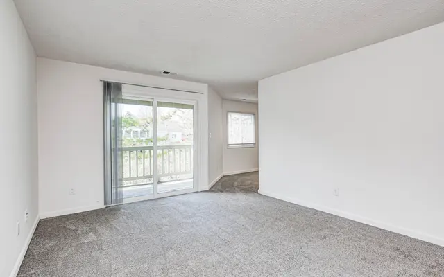 A spacious empty living room with carpeted flooring, featuring sliding glass doors leading to a balcony, and another window on the adjacent wall allowing natural light to enter.