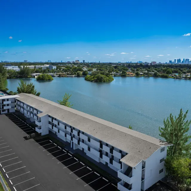 An aerial view of a modern low-rise building with a parking lot situated alongside a calm lake, surrounded by lush greenery and city skyline in the distance.
