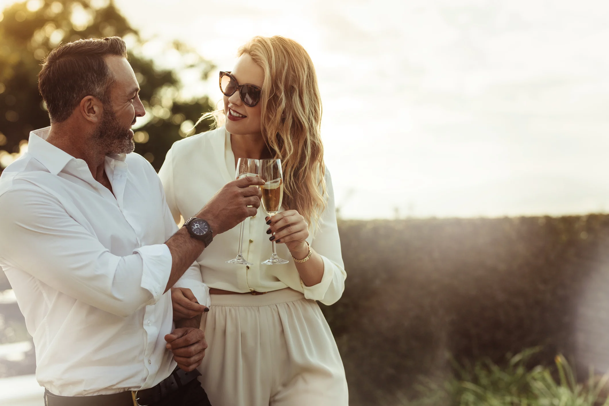 A man and a woman dressed elegantly are enjoying a toast with champagne glasses in an outdoor setting.