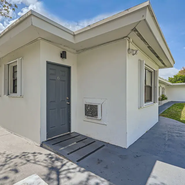 Exterior view of a modern single-story house with a grey door and air conditioning unit on the side, surrounded by a grassy area and blue sky in the background.