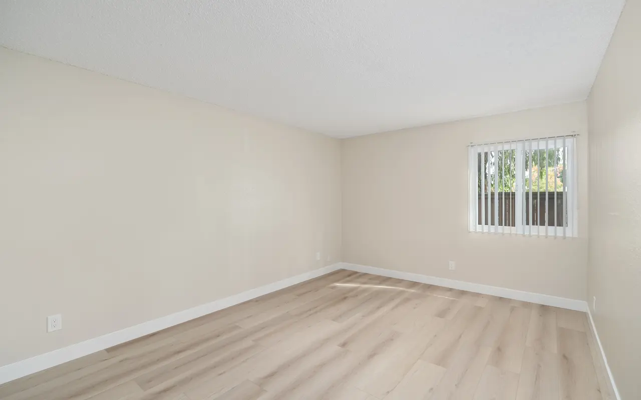 An empty room with light-colored walls and a wooden floor, featuring a window with blinds.