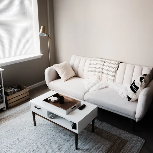 Cozy living room with a white couch, coffee table, and warm lighting, featuring a window with blinds and decorative elements.