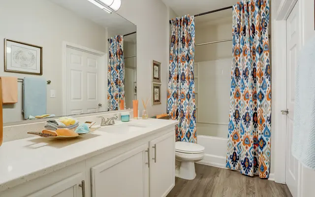 A bright modern bathroom featuring a double vanity with a white countertop, colorful patterned shower curtains, and a bathtub behind the curtains.