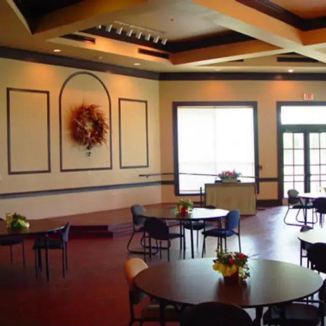 Interior view of a spacious event room featuring round tables with floral centerpieces, a small stage, and large windows providing natural light.