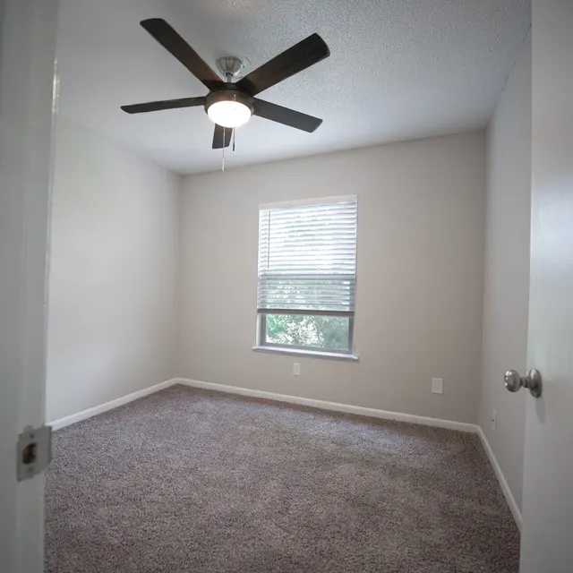 A vacant room featuring light colored walls and a ceiling fan. The room has carpeted flooring and a window with blinds, showing trees outside. A door is partially open, revealing part of the entryway.