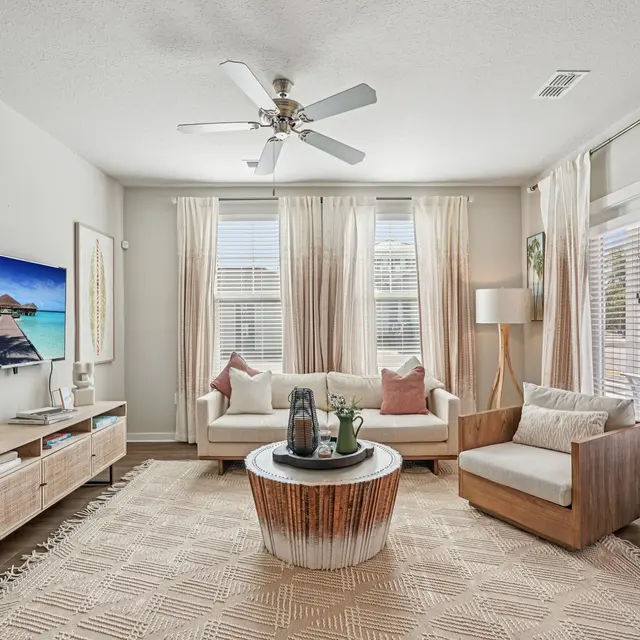 Modern Coastal Living Room A bright and airy living room featuring a white sofa and armchair, light drapes, and a television displaying a tropical beach scene. The room includes a wooden coffee table and a textured area rug on the floor.